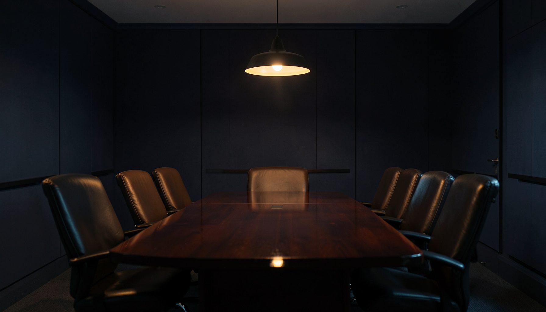 A dimly lit conference room with a long polished table and leather chairs, warm amber light spilling from a single pendant