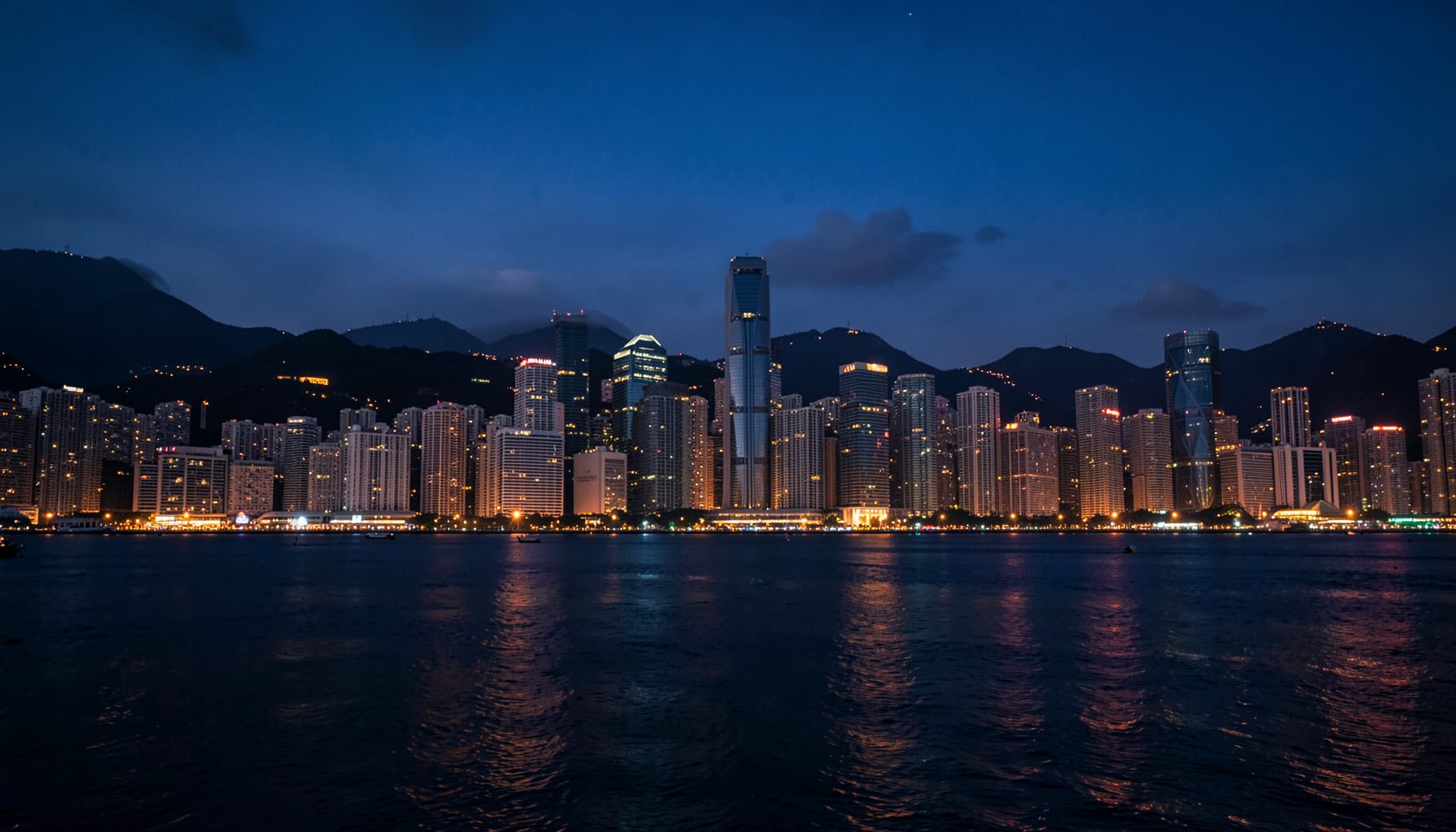 A moody skyline of Hong Kong at dusk with warm amber lights reflecting off the harbor, deep blue tones in the sky