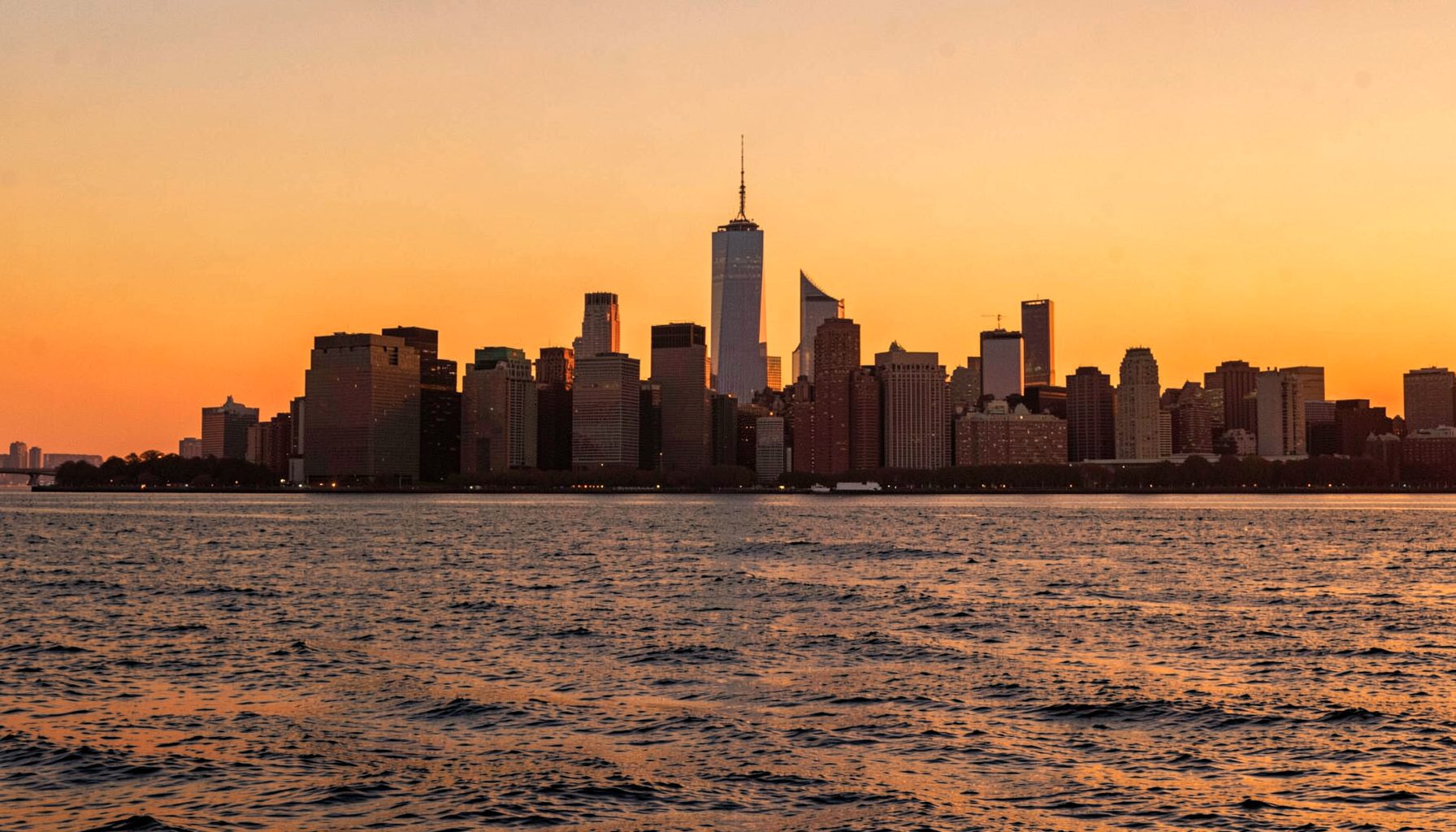 New York City skyline from across the water at golden hour, warm orange and amber tones, iconic silhouette